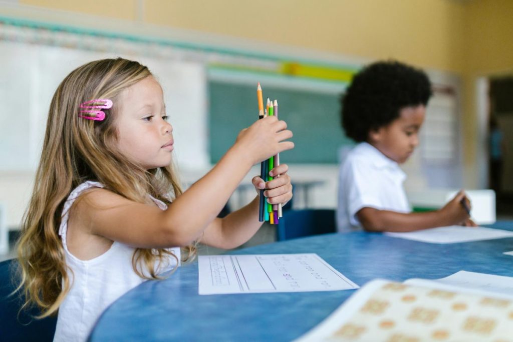 menina segurando varias cores de lapís na mão dentro da sala de aula